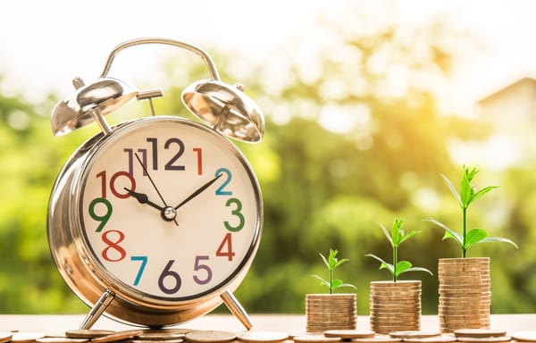 An alarm clock next to a stack of coins with a plant growing out of it.